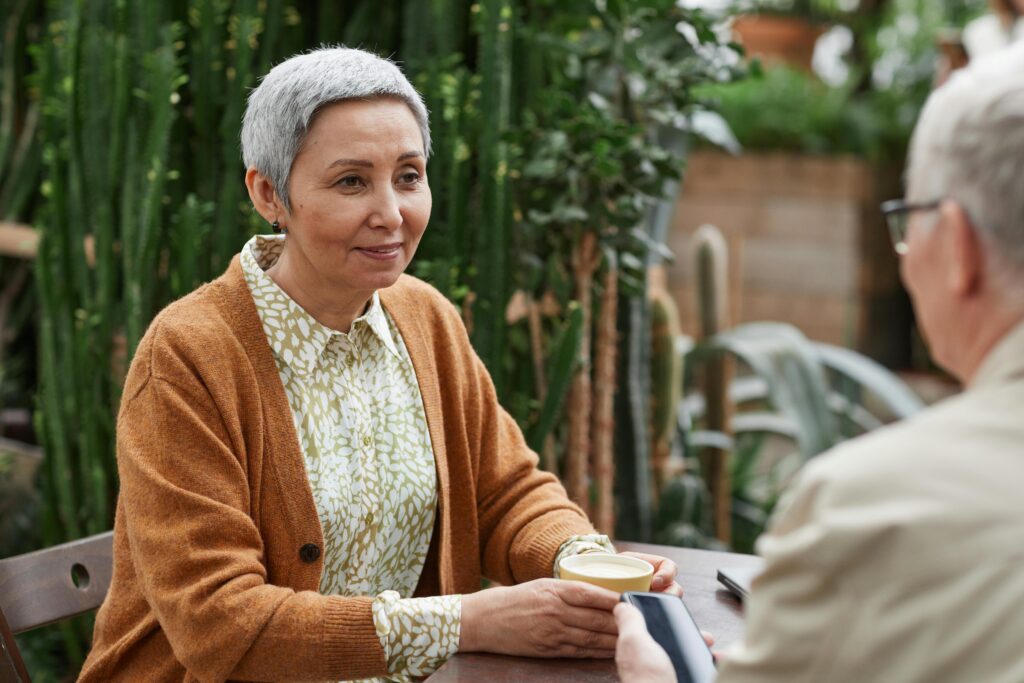 An older woman with short gray hair, wearing a brown cardigan, sits at a table holding a cup of coffee and smiles while talking to an older man in a garden or greenhouse setting, enjoying the peaceful atmosphere created by RestoRx.